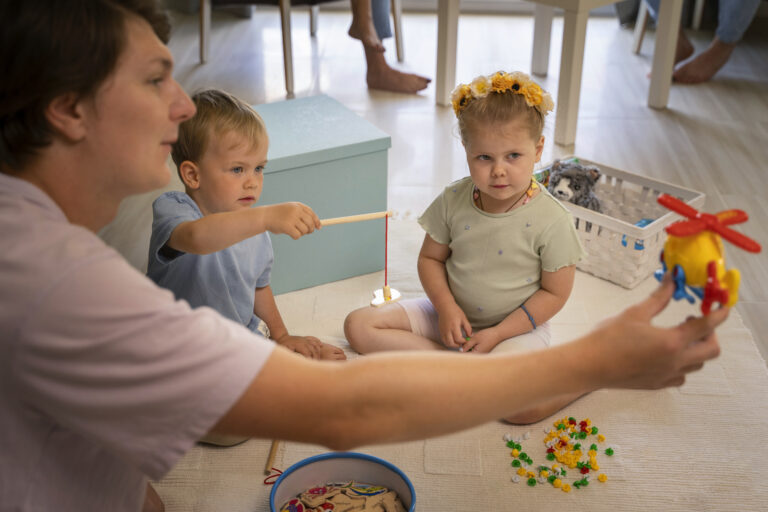 Kobieta bawi się z dwójką małych dzieci na podłodze, używając kolorowych zabawek edukacyjnych w jasnym pokoju.