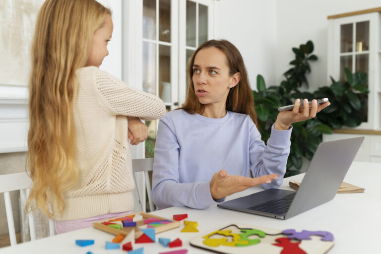 Kobieta siedzi przy biurku z laptopem i rozmawia z małą dziewczynką, na stole rozłożone kolorowe drewniane puzzle edukacyjne.
