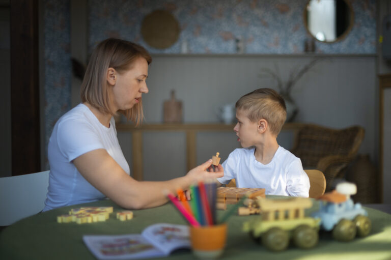 Kobieta siedzi przy stole z chłopcem i pracuje z nim przy użyciu drewnianych klocków edukacyjnych i kolorowych kredek.